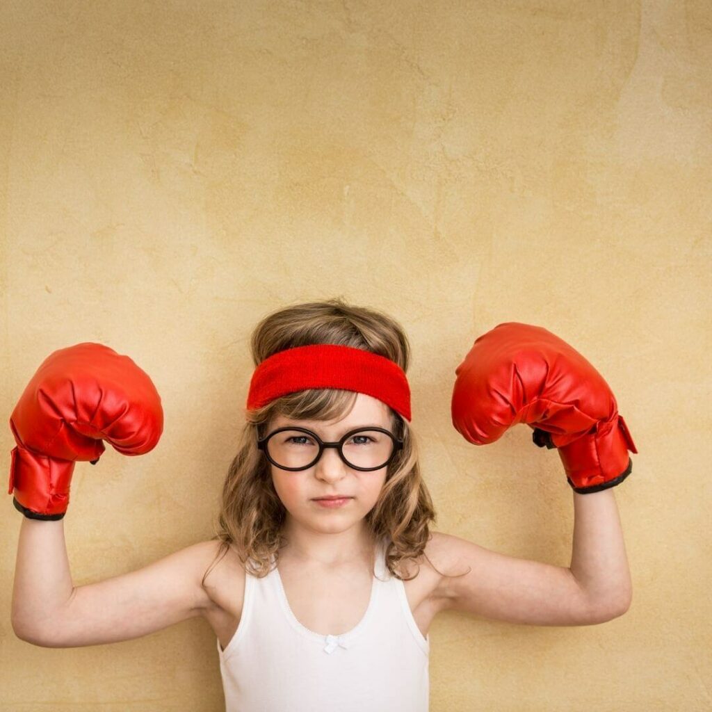 Das Bild zeigt ein kleines Mädchen mit blonden Haaren, schwarzer Brille und einem weißen Hemd. Sie trägt an den Händen rote Boxerhandschuhe und ein rotes Stirnband. Sie hat die Arme in Siegerpose gebracht und den Mundwinkel leicht hochgezogen.