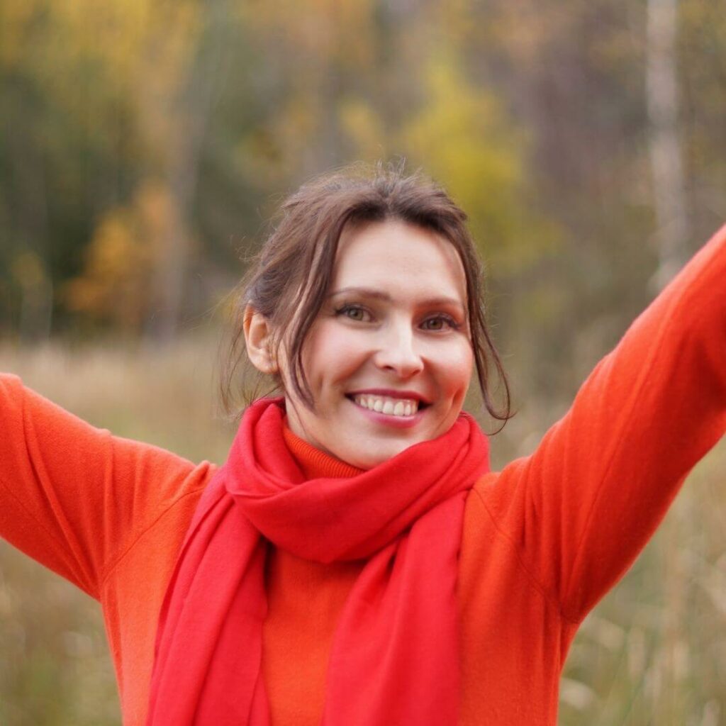 Das Bild zeigt eine junge Frau in einem roten Pullover. Sie hat die Arme in den Himmel gestreckt und lächelt in die Kamera. Sie strahlt Optimismus aus.